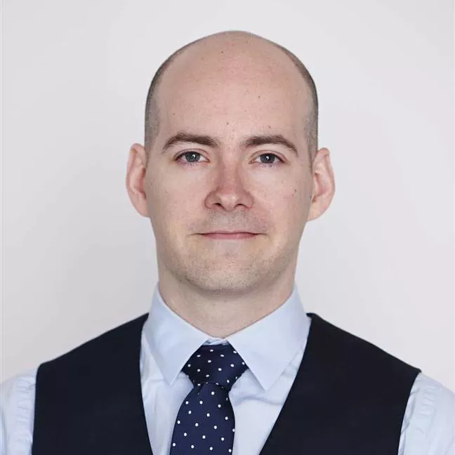 A man dressed in formal attire featuring a light blue shirt, a navy polka dot tie, and a dark vest. The background is plain white, emphasizing the professional and polished look. The outfit suggests a business or formal setting.